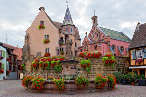 Charmante place du village de Niedermorschwihr avec ses bâtiments médiévaux colorés, jardinières de fleurs rouges et fontaine ornée