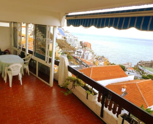 Terrace with sea view overlooking Santa Cruz de Tenerife coastline, white chairs, blue awning, ocean vista beyond apartment