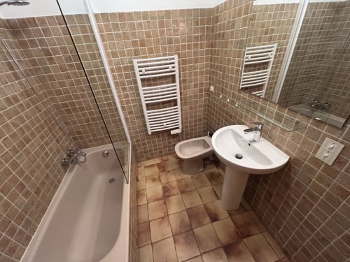 Modern bathroom with brown tile walls, white pedestal sink, bidet, bathtub with glass shower enclosure, and heated towel
