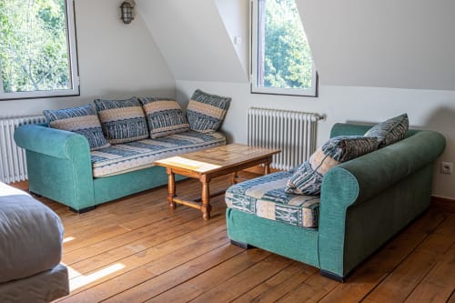 Lumineux salon mansardé avec sièges turquoise, coussins à motifs, table en bois, radiateur et verrières donnant sur les arbres
