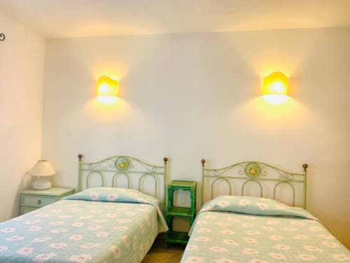 Camera doppia con letti in metallo verde, applique gialle e biancheria color crema presso l'Hotel Domu Incantada a Muravera