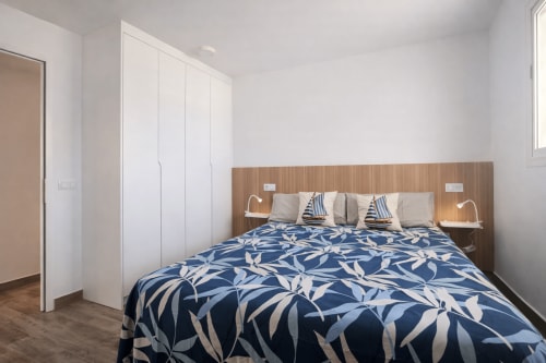 Modern bedroom with blue patterned bedspread, wooden headboard, white wardrobe, and natural light in La Aldea apartment