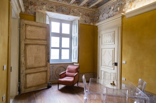 Camera elegante a Palazzo Fauzone con pareti ocra, soffitto ornato, porte in legno, finestra e poltrona rossa