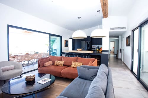 Modern villa living room with orange sofa, gray armchair, pendant lights, kitchen with dark cabinetry, and ocean view terrace