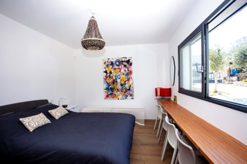 Modern bedroom at Locavilla villa in Colombes with navy bed, wooden desk, colorful artwork, and large window overlooking