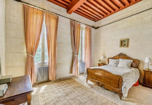 Chambre spacieuse à la Conciergerie Maurin Arles avec lit en bois, rideaux rayés, poutres apparentes et vue sur le jardin