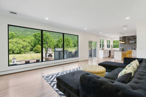 Modern mountain view living room with black sectional sofa, yellow ottoman, and expansive windows overlooking Highland Mills