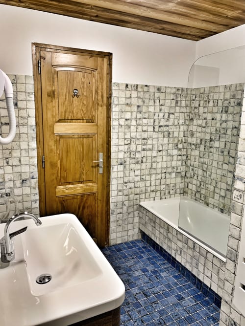 Bathroom in Chez Francine apartment with white sink, wooden door, tiled walls, bathtub and blue floor tiles