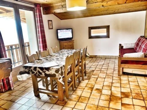 Spacious apartment dining area with wooden table, mountain lodge beams, TV, and balcony access at Chez Francine