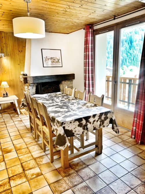 Dining room in Chez Francine apartment with wooden beams, fireplace, cow-print tablecloth, and mountain views through large