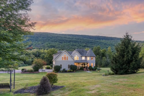 Illuminated farmhouse villa with mountain views in Highland Mills, nestled in lush valley landscape at sunset.