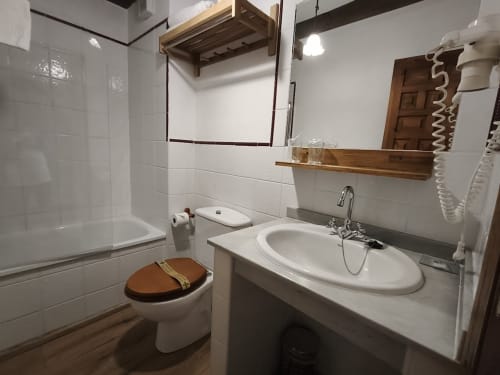 Clean bathroom with white tiles, round sink, wooden mirror shelf, toilet with wood seat, and shower at Posada La Colodra
