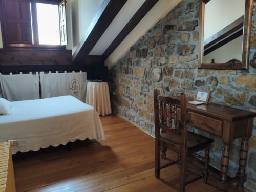 Rustic attic bedroom at Posada La Colodra with stone wall, wooden beams, bed, desk, and mountain views in Spain