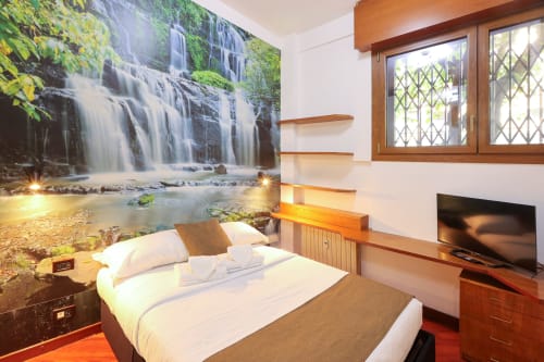 Accogliente camera studio all'hotel Solumnia di Milano con murale cascata, scaffali in legno, letto, TV e finestre con vista sulla città