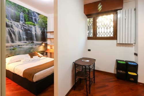 Studio Solumnia Milano con camera da letto, opera d'arte cascata, letto in legno e zona pranzo con tavolo e cestini
