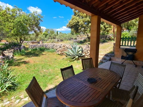 Wooden dining table with chairs on a covered terrace overlooking gardens at Domaine de L'Échappée Belle in Entrecasteaux