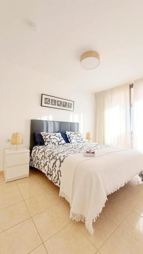 Bright bedroom with gray upholstered bed, patterned bedding, white nightstand with lamp, wooden ceiling light, framed art,