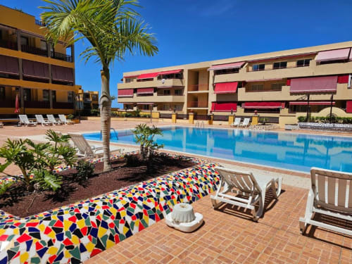 Resort pool with colorful mosaic lounge path, palm tree, and modern apartment buildings in Tenerife