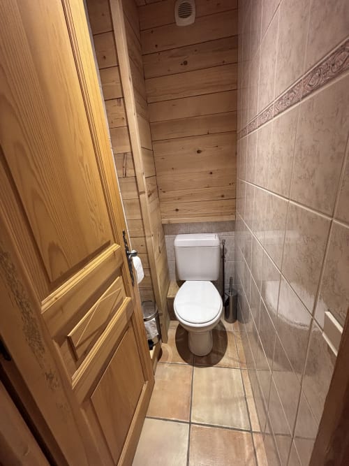 Compact bathroom in Chez Marion apartment with white toilet, beige tile walls, wooden ceiling, and wooden door in Notre Dame