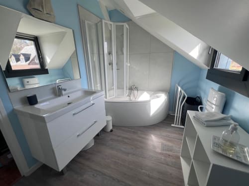 Salle de bain mansardée moderne à Destination Etretat avec lavabo blanc, baignoire d'angle, douche, murs bleus et puits de lumière