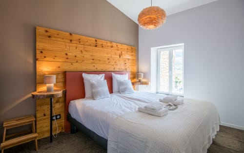 Chambre moderne dans un appartement à Millau avec tête de lit en bois, coussin rouge, draps blancs, lampe suspendue et vue sur la ville