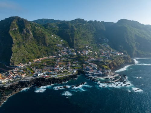 Vista aérea da aldeia costeira de Funchal com casarões coloridos, jardins em socalcos e ondas turquesas do oceano