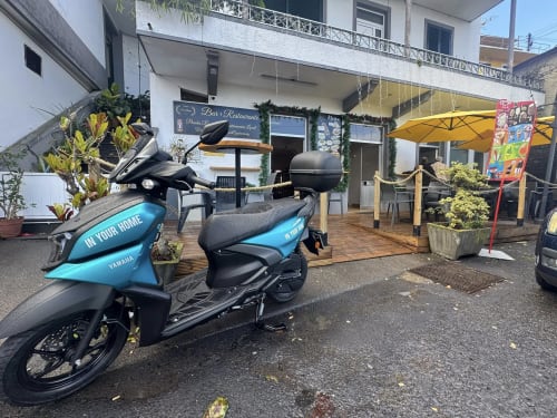 Scooters Yamaha azuis estacionadas no pátio de SpecialStays Funchal com restaurante, guarda-sóis amarelos e plantas.