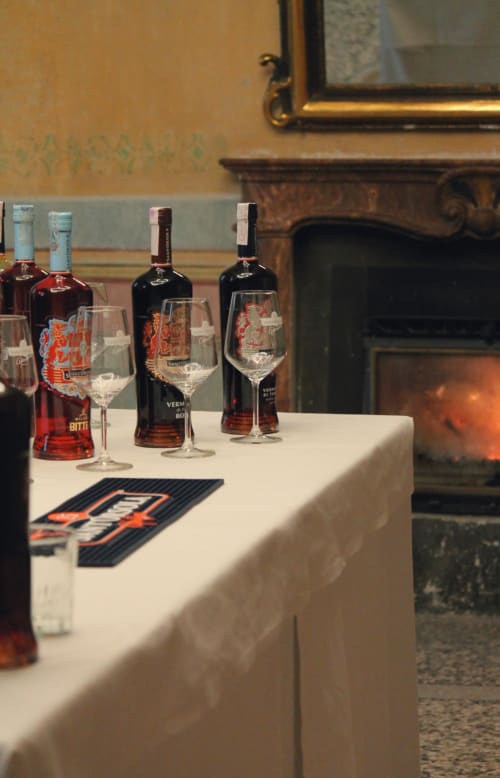 Bottiglie di vermouth e bicchieri brandizzati esposti su tavolo con tovaglia bianca al Palazzo Fauzone Relais & Spa a Mondovì, Italia