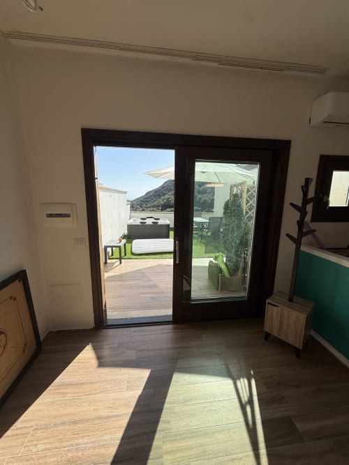 Moderno interno di appartamento con porte in vetro e cornice scura che si aprono su terrazza in legno, vista montagne e mare in Sardegna