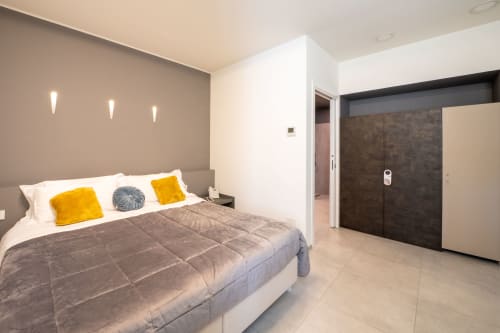 Camera da letto moderna e accessibile dell'Hotel Belvedere Resort con letto grigio, cuscini gialli, armadio scuro e applique