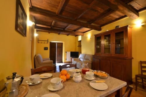 Sala da pranzo giallo brillante con travi in legno, tavolo preparato con arance, salotto con divano e TV all'Agriturismo Buriano