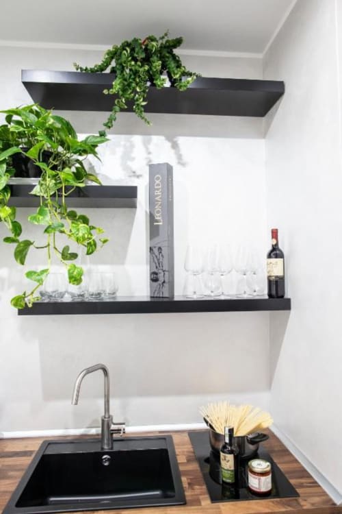 Elegante scaffalatura nera con piante in vaso, bicchieri, bottiglia di vino e scatola Leonardo sopra il lavello della cucina a Domus Gaudia
