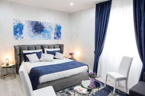 Elegante camera da letto di Domus Gaudia Roma con letto imbottito grigio, accenti blu navy, biancheria bianca e arte moderna