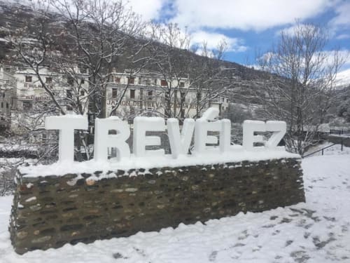 Trevélez town sign with snow-covered stone base, snow-covered landscape, bare trees, and white buildings nestled in mountains