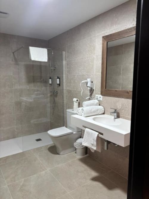 Baño moderno del Hotel Mulhacén Trevélez con ducha de cristal, lavabo blanco, inodoro y toallas enrolladas en Granada