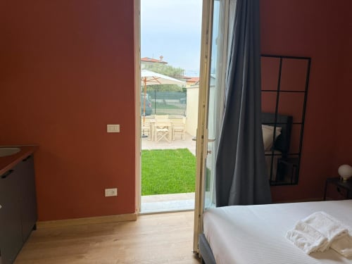 Camera da letto moderna con pareti rosse, letto bianco e porta in vetro che si apre sul giardino verde dell'appartamento Mistral Holidays a Marina di