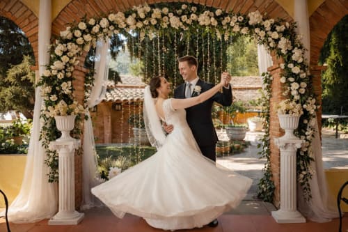 Pareja de novios bailando bajo un arco de ladrillo decorado con flores y columnas blancas en una terraza exterior.