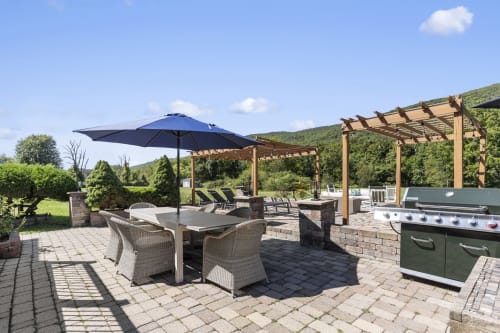 Luxury outdoor patio at Malkita Homes with dining area, blue umbrella, pergola, and built-in grill overlooking Highland Mills