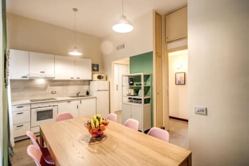 Luminosa zona cucina-pranzo in appartamento a Roma con mobili bianchi, tavolo in legno, sedie rosa e parete verde accento