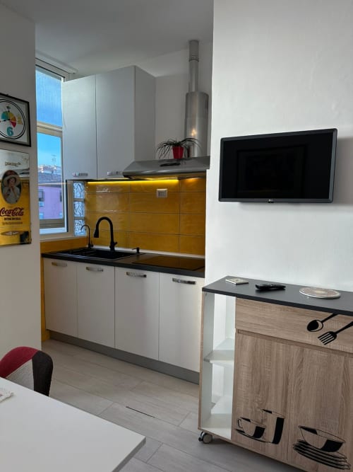 Modern kitchen in Aparthotel Lumi, Catania with yellow backsplash, white cabinetry, stainless steel hood, and wall-mounted TV