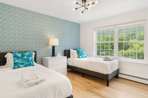 Bright twin bedroom at Malkita Homes with turquoise patterned wallpaper, white bedding, blue pillows, and large windows