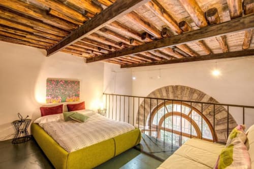 Camera da letto in loft moderno a Roma con travi di legno a vista, letto giallo, finestra ad arco e soppalco