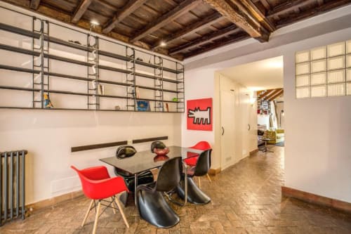 Zona pranzo di moderno appartamento romano con sedie rosse, tavolo nero, travi in legno a vista e scaffalature metalliche