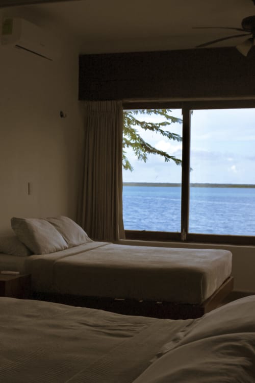Habitación frente al lago en Tucan Village Bacalar con vistas al lago a través de puertas de cristal, ropa de cama blanca y escenario de playa
