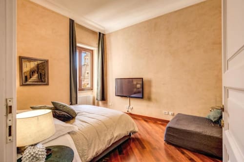 Camera da letto spaziosa in appartamento romano con pareti calde, pavimenti in legno, letto, TV e vista su monumento dalla finestra