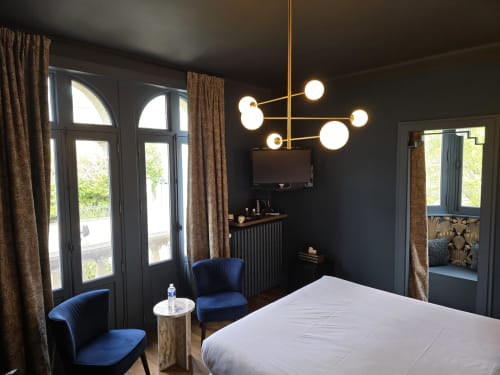 Chambre moderne au Castel Bayeux avec murs sombres, fauteuils en velours bleu, luminaire suspendu doré et vue sur la cathédrale