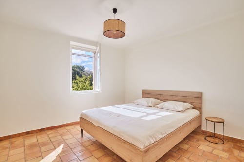 Chambre lumineuse avec lit en bois, suspension en rotin, sol en terre cuite et vue sur le jardin à la villa CIRELINE à Rognes