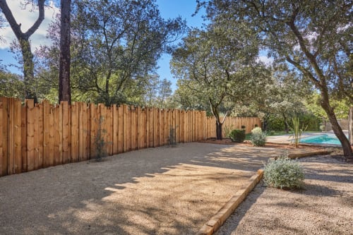 Jardin privatif de la villa CIRELINE à Rognes avec clôture en bois, terrasse en gravier, arbres matures et aperçu de la piscine