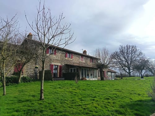 Stone farmhouse with red shutters at Domaine de Beau Site B&B in Peyrat-de-Bellac, surrounded by green lawns and bare trees.