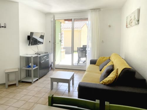 Studio moderne au Vallon des Sources avec canapé gris, coussins jaunes, TV, kitchenette et accès à la terrasse